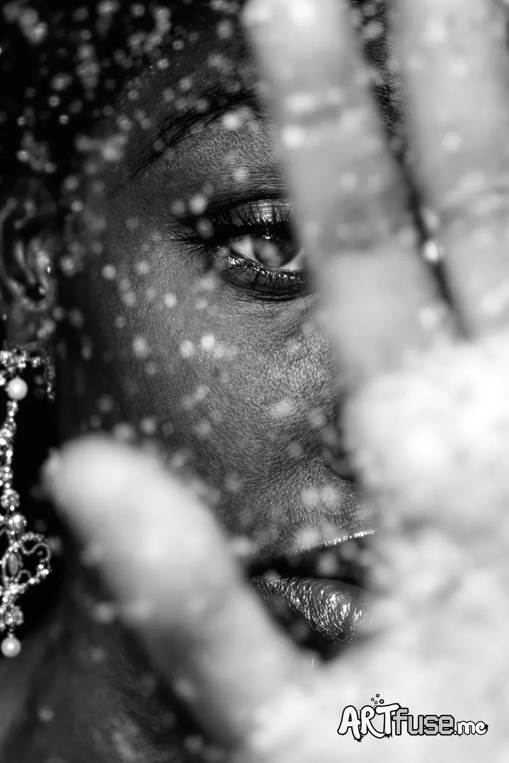 A close-up, black and white portrait of a person of African descent, likely in their late 20s or early 30s. The face is partially obscured by a hand or veil, the person is facing the viewer at a slight angle. Water droplets, covering the surface in front of the subject, add a layer of texture and blurred distortion. The person’s eye is open and expressive, conveying a sense of introspection. They have full lips and a defined nose. The subject is wearing a decorative earring with intricate detail