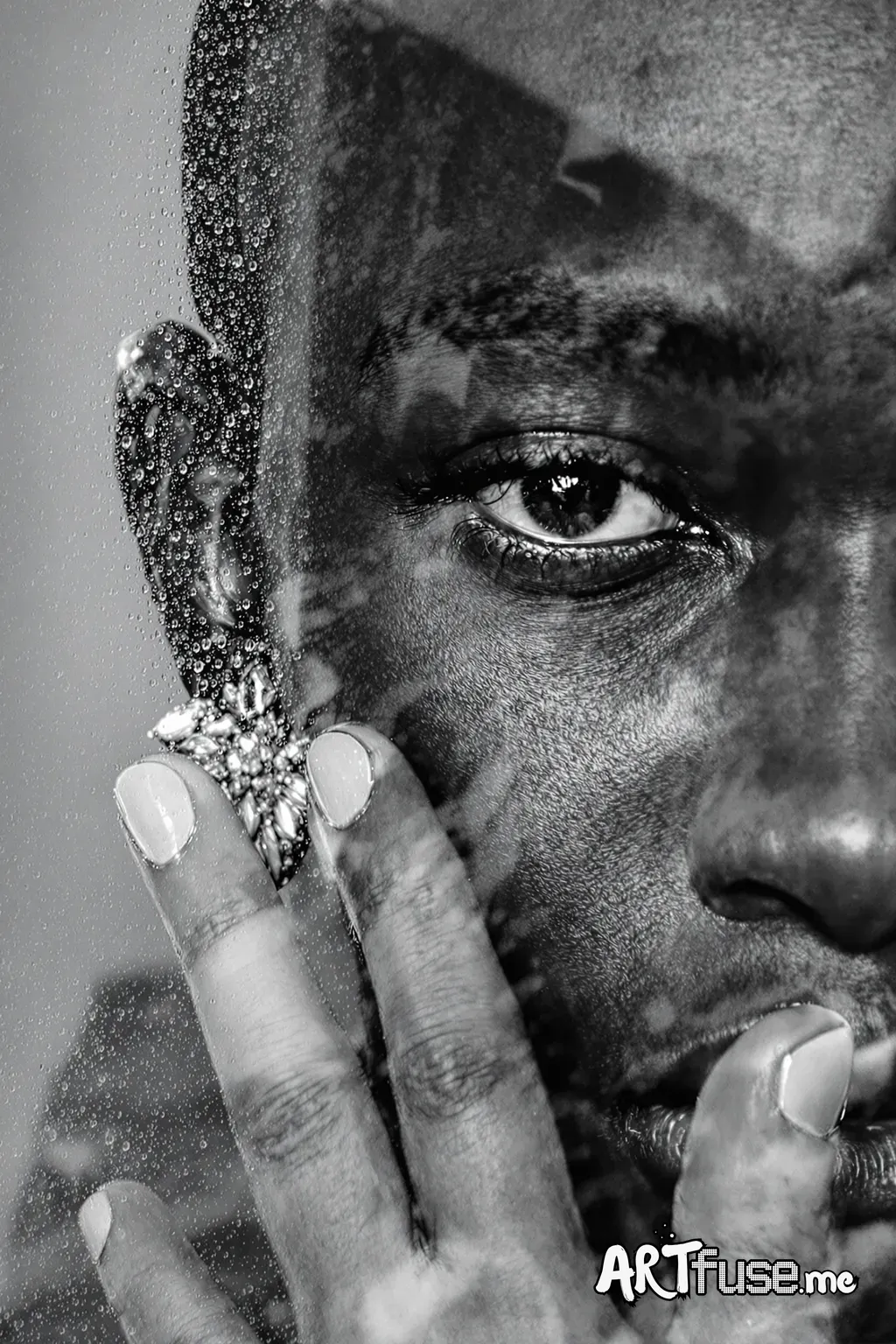A close-up, monochromatic portrait of a Black individual, man face partially obscured by a sheet of glass covered in droplets of water. The subject, likely in their late 20s or early 30s, has smooth, dark skin with visible texture, and a bald head. One eye is visible, gazing directly towards the viewer with a piercing intensity. The subject wears an ornate earring adorned with what appears to be jewels, adding a touch of elegance. A hand, likely their own, is gently resting behind the glass, b